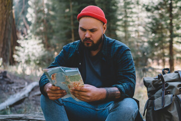 Young male traveler concentrated on choosing route for hiking sitting on old tree near backpack in forest, hipster guy wanderlust reading map checking location and destination exploring wild landscape