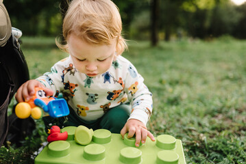 Child 2 year old play constructor game sitting in green grass in park on sunny summer day. Outdoors...