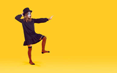 Full length portrait of funny woman in halloween costume short black dress, striped knee socks and black hat with scary mad makeup. She is dancing and showing arm to copy space on yellow background.