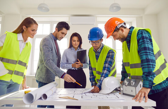 Construction team having discussion at work meeting. Group of multiracial male and female architects and builders wearing safety hard hats standing around office table looking at paper blueprint plans - Powered by Adobe