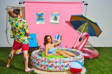 Young man spraying water from hose at beautiful woman in vibrant kiddie pool. Fun and festive summer vibe. Concept of pop art, remote work, travelling, party, recreation, lifestyle, fashion and style.