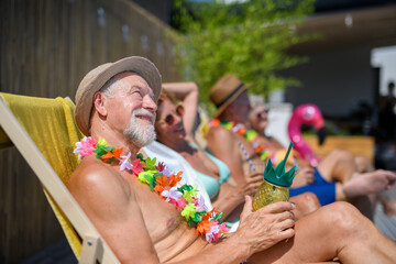 Group of cheerful seniors sitting lounge chairs by a swimming pool and sunbathing. Handsome senior man enjoying a hot day outdoors in backyard with friends.