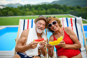 Beautiful elderly couple enjoying their vacation. Seniors having romantic time, sitting by swimming pool in lounche chairs sunbathing.