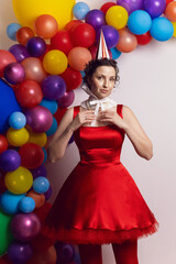 Funny clown woman in a red dress and a hat stands against a background of colored balloons