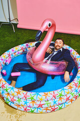 Young office worker in formal suit relaxing in kiddies swimming pool on flamingo inflatable-ring...