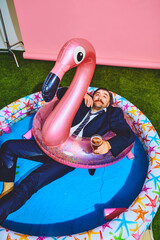Dreaming businessman in formal suit relaxing in kiddies swimming pool on flamingo inflatable-ring...