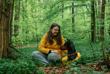 young woman walking with her doberman dog in the forest. Wearing yellow rain jackets girl and dog playing outdoors. Human and dog friendship concept