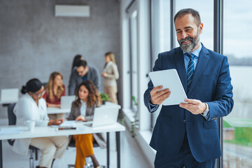 Happy middle aged business man ceo wearing suit standing in office using digital tablet. Smiling mature businessman professional executive manager looking away thinking working on tech device.
