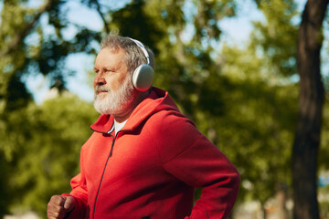Senior man jogging in park, in red hoodie and headphones. Early morning running routine. Outdoor training. Concept of sport, aging, active and healthy lifestyle, health care