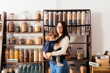 Smiling Mother Holding Her Baby In A Sustainable Bulk Food Store and Looking at Camera