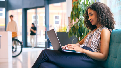 Businesswoman Sitting In Lobby Of Modern Office Building Working On Laptop