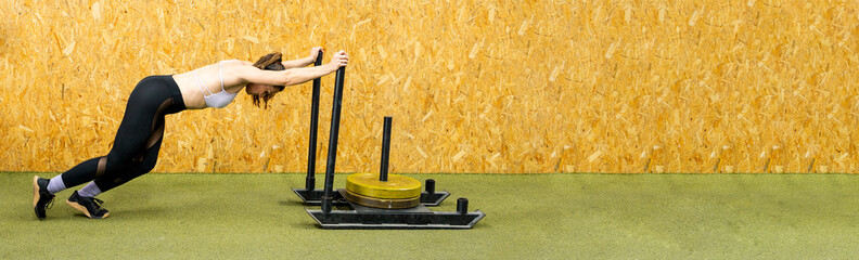 A middle-aged adult woman performs physical exercise inside a gym.The 50-year-old pushes a dragging...