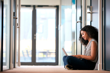 Businesswoman Sitting On Floor In Corridor Of Modern Office Building Working On Laptop