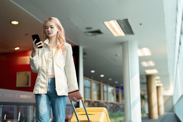 Young female traveler with suitcase using smartphone at airport. Modern travel, casual outfit, waiting for flight