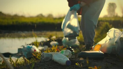 On the shore of a pond, a young man collects plastic waste, carefully sorting it. It places plastic bottles and other waste in a trash bag to ensure they are recycled and disposed of sustainably