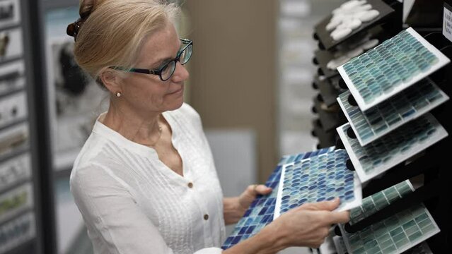 Happy mature woman touching glass wall tiles and swimming pool tiles while shopping through samples for home redecorating renovation project, in a design center store.