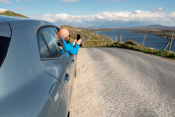 Car parked off small road, male driver taking picture on his smart phone of beautiful scenery, Sky...