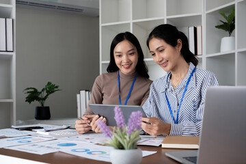 Two Asian businesswomen discussing work using a tablet in a modern office. Concept of teamwork and professional collaboration