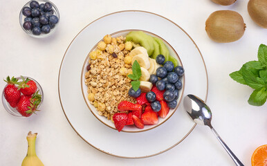Granola with strawberries, kiwi, banana and blueberries in a round plate on a white table.