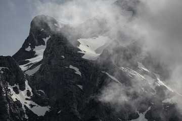 Alpine peaks rise through clouds, their jagged summits shrouded in mist. The scene captures the ethereal and majestic beauty of the towering mountains in soft, diffused light.