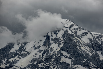Alpine peaks rise through clouds, their jagged summits shrouded in mist. The scene captures the ethereal and majestic beauty of the towering mountains in soft, diffused light.