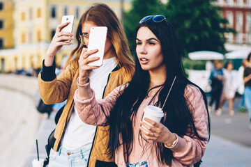 Two female tourists with coffee in hand walking in new town and making photo on modern telephone...