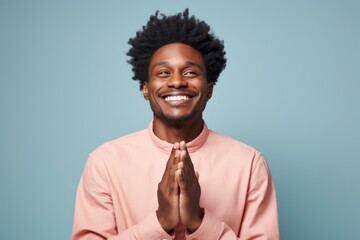 Fototapeta premium Portrait of a tender afro-american man in his 20s joining palms in a gesture of gratitude on solid color backdrop