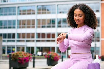 Woman Wearing Fitness Clothing Sitting Outdoors Checking App On Fitness Monitor Or Smart Watch