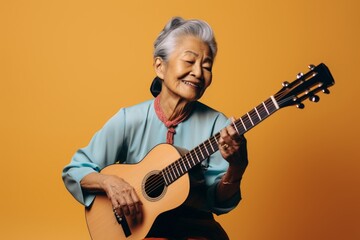 Obraz premium Portrait of a tender asian woman in her 80s playing the guitar while standing against solid color backdrop