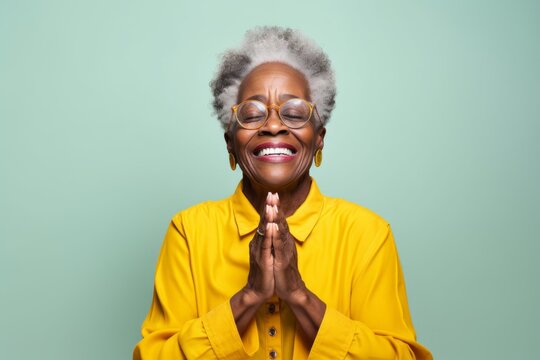Portrait of a grinning afro-american elderly woman in her 90s joining palms in a gesture of gratitude isolated in solid color backdrop - Powered by Adobe