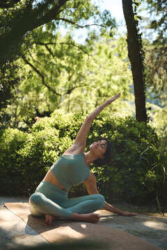 Happy mindful young Hispanic woman meditating doing yoga breathing exercises feeling peace of mind, mental balance sitting in lotus pose stretching in green nature park outdoors. Vertical.