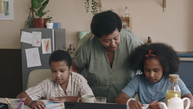 Medium of elderly African American woman with short hair hugging and stroking two junior grandchildren, who are sitting at kitchen table and drawing with colour pencils after school