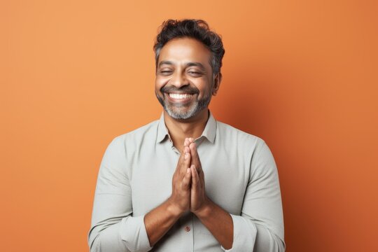 Portrait of a joyful indian man in his 40s joining palms in a gesture of gratitude in front of solid pastel color wall