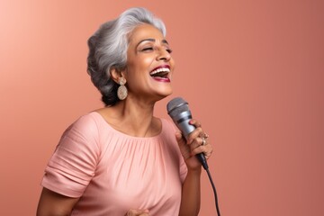 Portrait of a tender indian woman in her 50s dancing and singing song in microphone while standing against solid pastel color wall