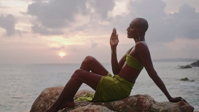 Divine non-binary black person in long open dress, brass jewelry poses on rocky hill top above dramatic ocean sunset skyline. Queer lgbtq fashion model in open outfit sits on a hill. Pantheon concept