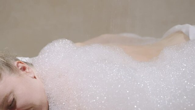 Spa worker washes woman under bubble soap foam with a stream of water in hammam shower. Female relax, gets body skin treatment, cleansing, wash. Anti aging and anti cellulite therapy in sauna.