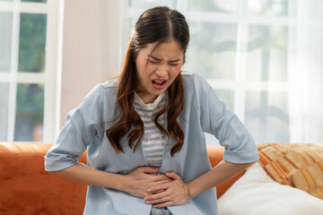 Woman experiencing stomach pain while sitting on the couch, expressing discomfort and holding her...