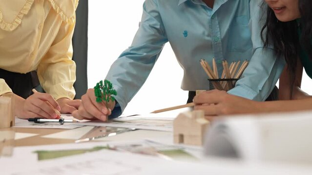 Architect and safety engineer discuss about position of tree use blueprint, model on table with architectural document, measurement tool and wooden block scatter around at modern office. Delineation.
