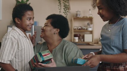 Medium zoom-in of two African American children happily running towards grandmother with gifts, congratulating on her birthday, embracing and kissing her with warmth and love - Powered by Adobe