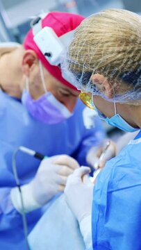 Doctor bent above the patient carefully performing the surgery on thyroid. Female nurse wearing yellow protective glasses assists at operation. Vertical video.