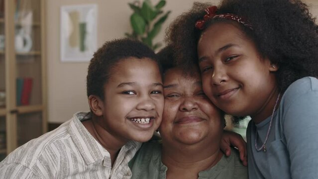 Portrait of two happy loving young African American siblings hugging grandmother with affection and smiling for camera, while posing together in kitchen