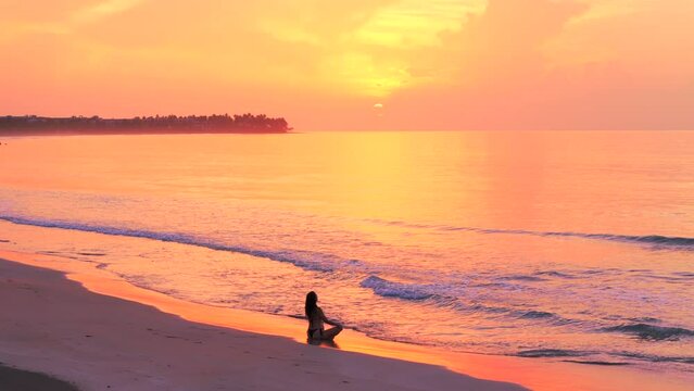 The silhouette of a young slender woman in yoga meditation pose sitting on a wild tropical beach with no people at beautiful sunset. Relaxation and calm to relieve stress and anxiety