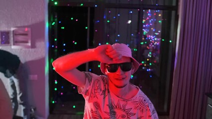 Young guy in panama hat and black sunglasses dances merrily to music, at party at night, among multi-colored flickering lights, he waves his arms and celebrates his birthday.