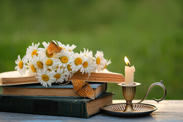 candlestick with burning candle, butterflies, old books and chamomile flowers on table in garden....