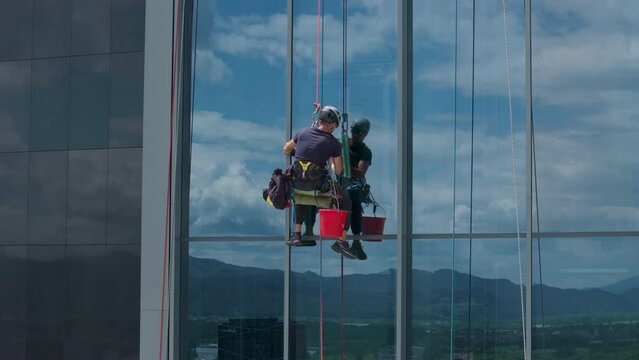 Aerial shot - High risk, dangerous job of a professional window cleaner. Window cleaner suspended on a rope cleaning window on a high rise building.