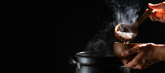 The cook is cooking rice porridge in a boiling clay pot with steam rising and using a wooden ladle...