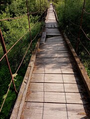 Fototapeta premium Old suspension bridge made of wood and metal cables over a river in a dilapidated condition with rotten planks and rusted metal structure. Dangerous emergency pedestrian bridge
