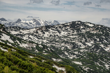 The view depicts a mountain towering over a valley, with lush greenery contrasting the rugged...