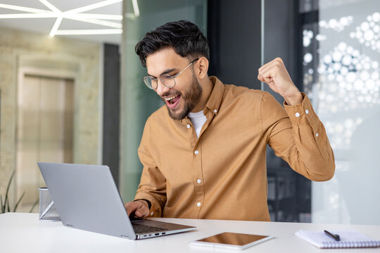 Happy young muslim man sitting in office at table, looking at laptop screen and enjoying achievement and success, making victory gesture with hand yes