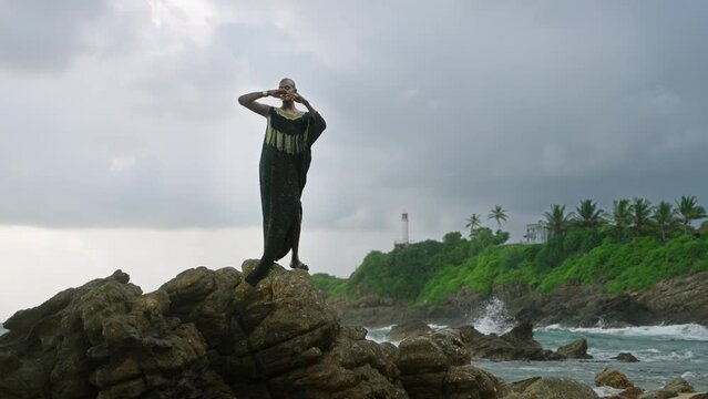 Godlike gender fluid person of color in luxury dress, jewelry stands on cliff peak at scenic ocean. Pride extravagant fashion model in epic outfit posing like goddess in tropics. Olymp concept.